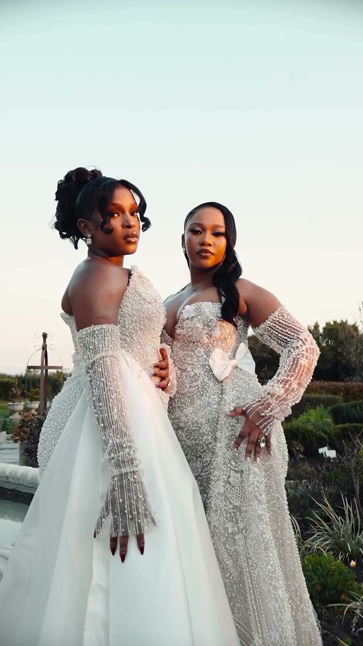 Two OZYDE bridal gowns side by side in a garden at dusk — a pearl gown with satin overskirt beside a crystal-beaded gown with a satin bow